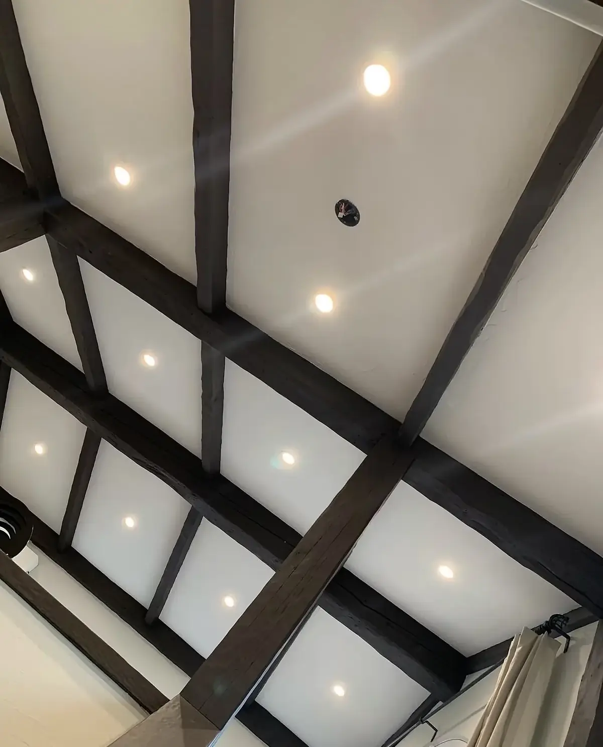 Sloped white ceiling divided into bays by dark decorative beams, with a round recessed light centred in each bay