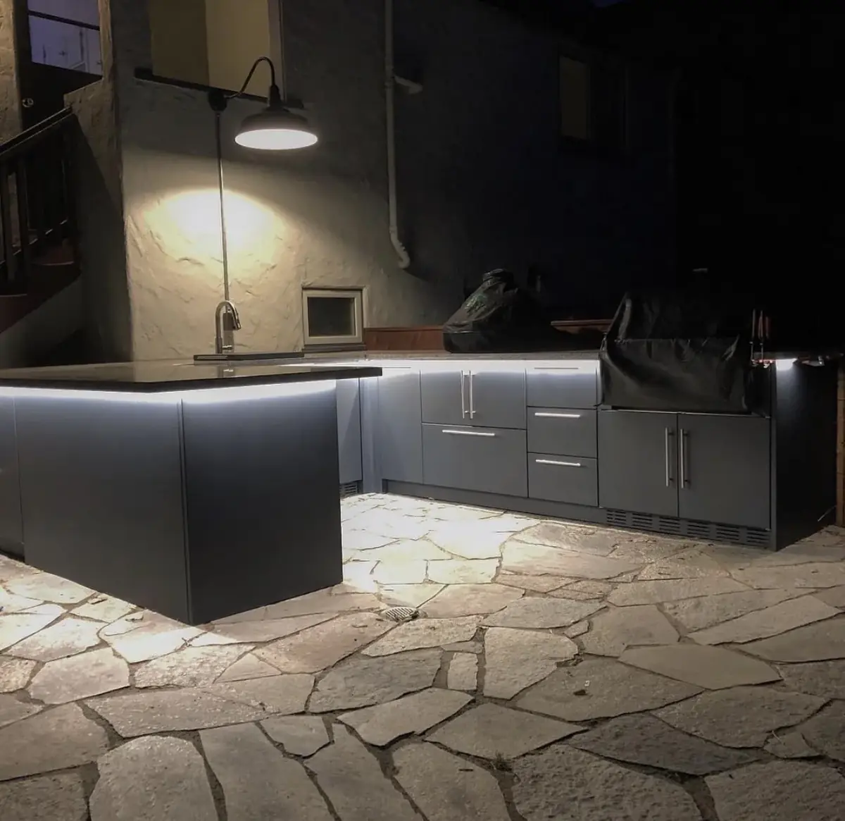 Outdoor kitchen at night with an LED strip glowing under the counter edge onto flagstone paving and a gooseneck wall light above