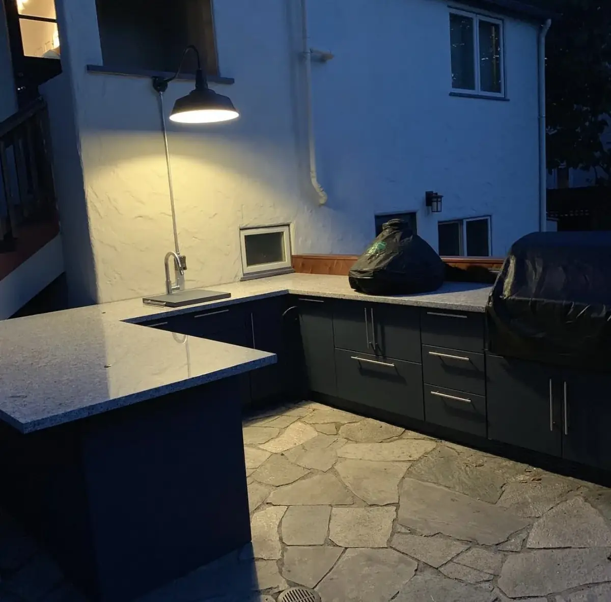 Outdoor kitchen at dusk with a granite counter, dark cabinets and a lit gooseneck wall light mounted on a white stucco wall