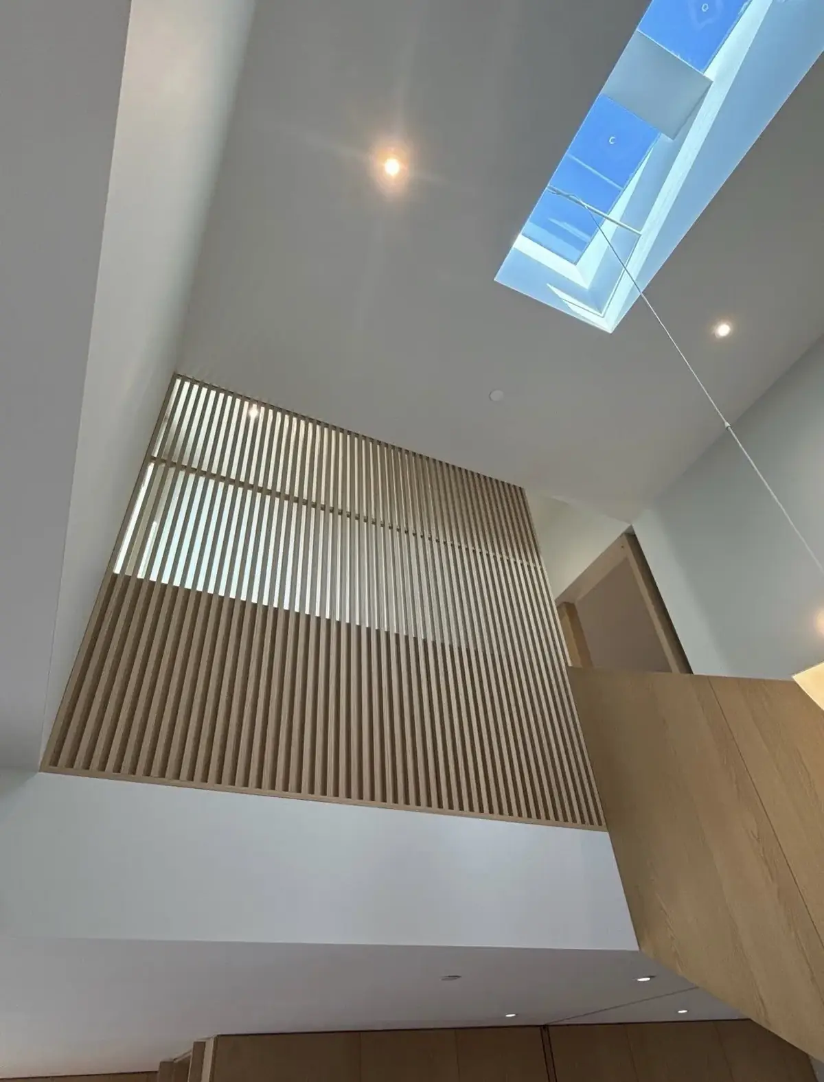 Double-height white hall with a long skylight, a recessed light beside it and a vertical oak slat screen below