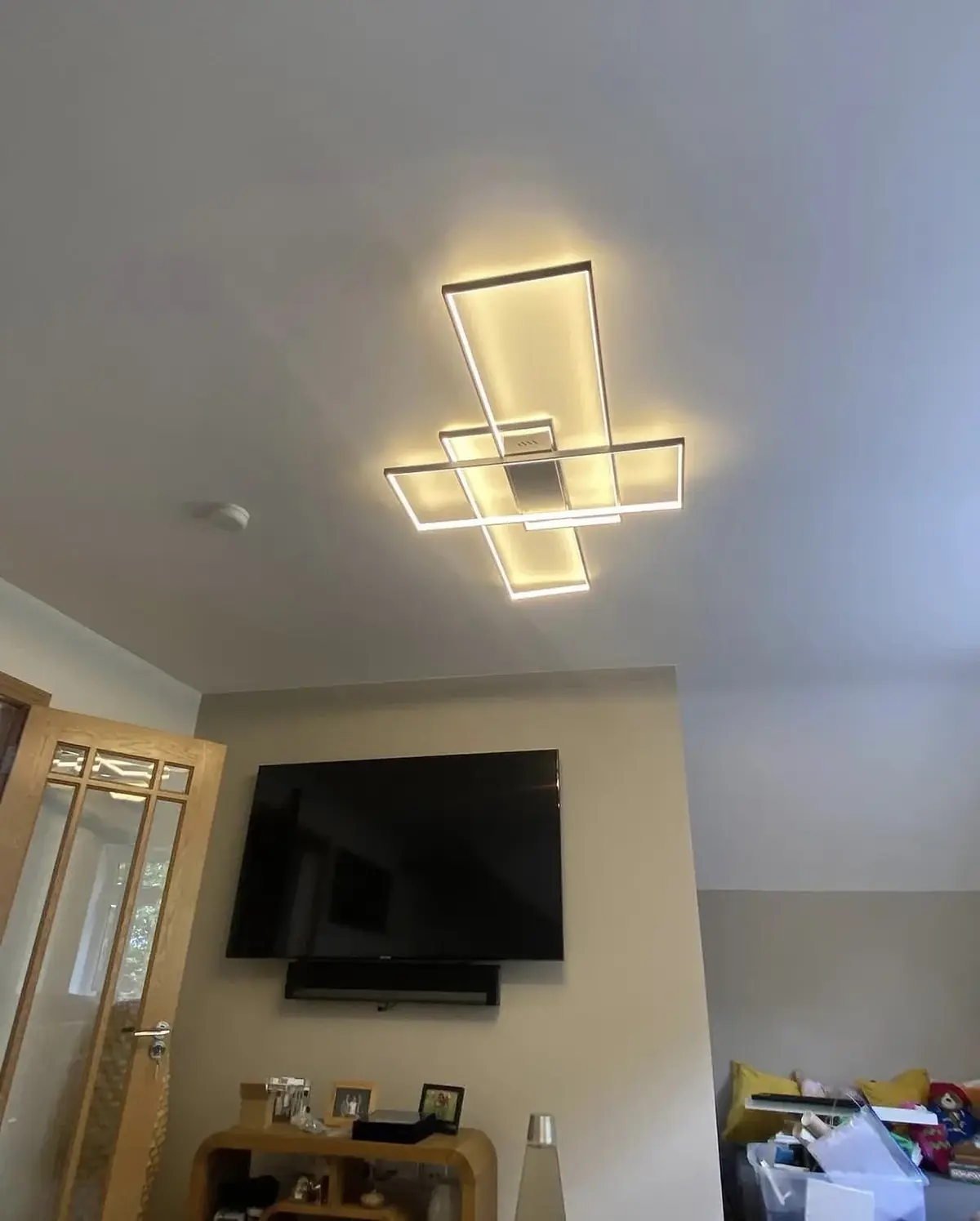 Surface-mounted LED ceiling fixture made of overlapping rectangular frames, lit, above a living room with a wall-mounted television
