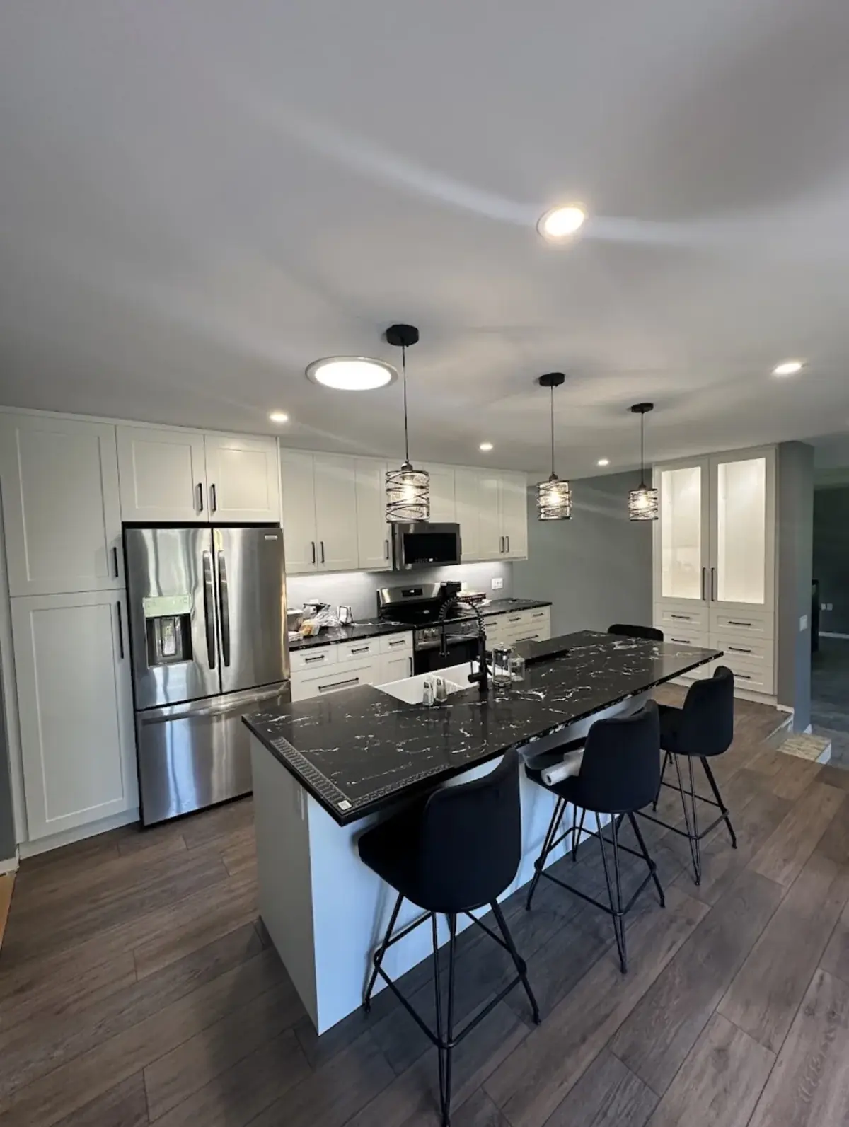 White kitchen with a black stone island, three pendant lights above it, recessed ceiling lights and dark bar stools