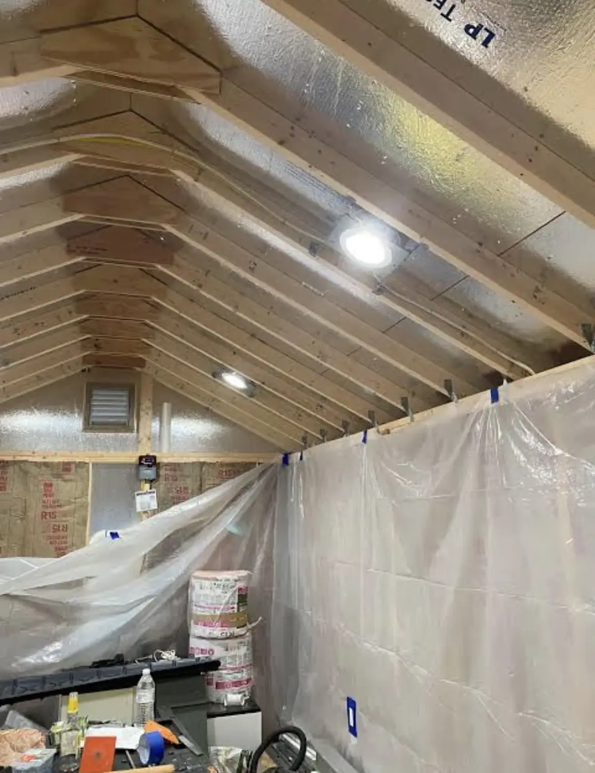 Vaulted loft ceiling with exposed rafters and foil-faced sheathing, two recessed LED lights switched on between the rafters, plastic sheeting and insulation bags below