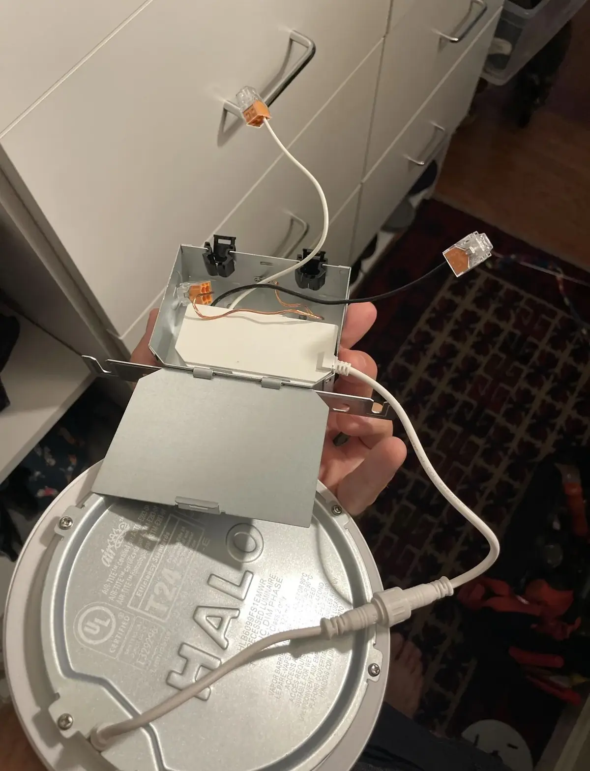 Hand holding a slim round recessed LED light with its metal junction box open, showing push-in wire connectors and the plug-in driver lead
